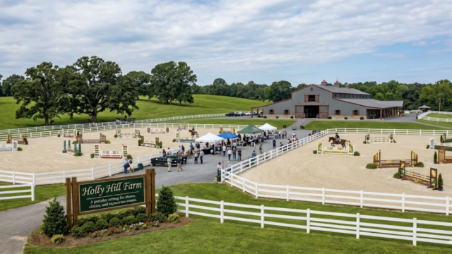 Horse show arena at Holly Hill Farm with jumps, tents, and a large barn surrounded by white fencing and green fields under a cloudy blue sky.